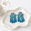 A pair of handmade clay earrings with a dalmatian-print circle top, teal arch middle, and five glittery teal teardrop dangles on a white ceramic dish.