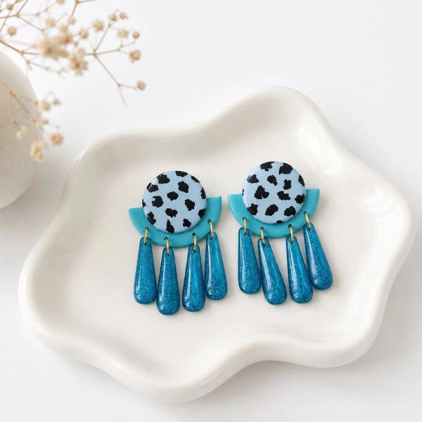 A pair of handmade clay earrings with a dalmatian-print circle top, teal arch middle, and five glittery teal teardrop dangles on a white ceramic dish.