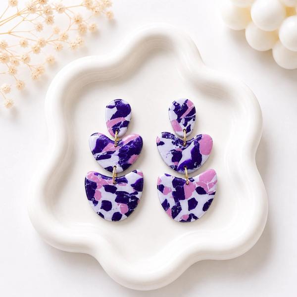 Three-tiered handmade clay dangle earrings with a white, purple, and pink terrazzo pattern connected by gold jump rings, displayed on a white ceramic tray.