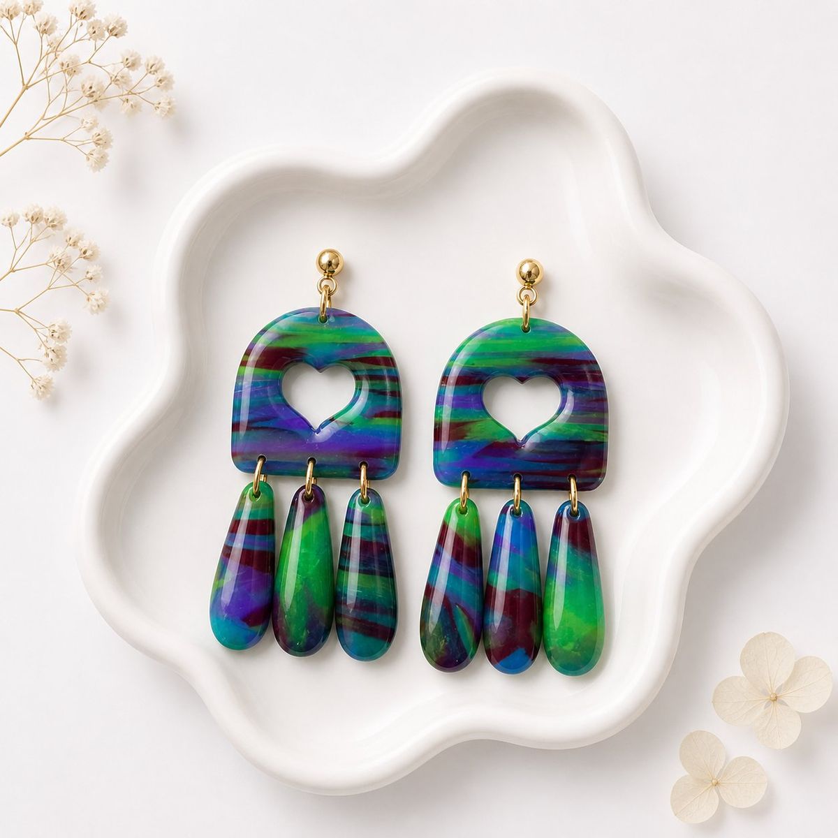 A pair of handmade clay dangle earrings with a marbled green, blue, purple, and burgundy arch top featuring a heart cutout, with three matching teardrop clay pieces hanging below, displayed on a white ceramic dish.