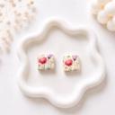 A pair of square clay stud earrings with a watercolour wildflower print in pink, purple, and yellow tones on a cream base, displayed on a white wavy ceramic tray.