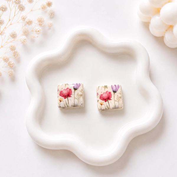 A pair of square clay stud earrings with a watercolour wildflower print in pink, purple, and yellow tones on a cream base, displayed on a white wavy ceramic tray.