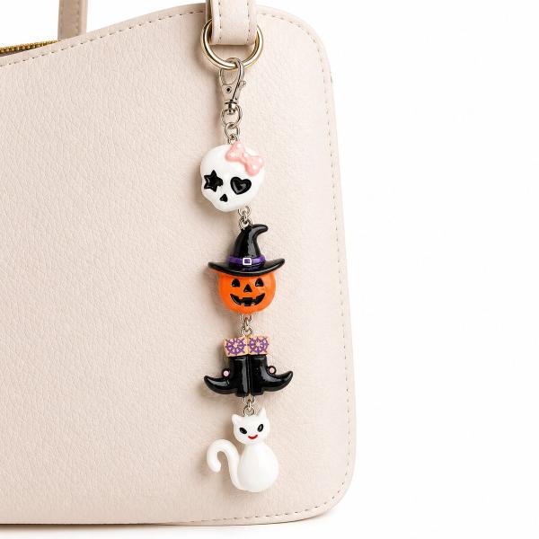 A Halloween keychain with four polymer clay charms — a skull with pink bow, a jack-o'-lantern wearing a witch hat, a witch figure with purple bow, and a white cat — hanging from a silver lobster clasp on a cream handbag.
