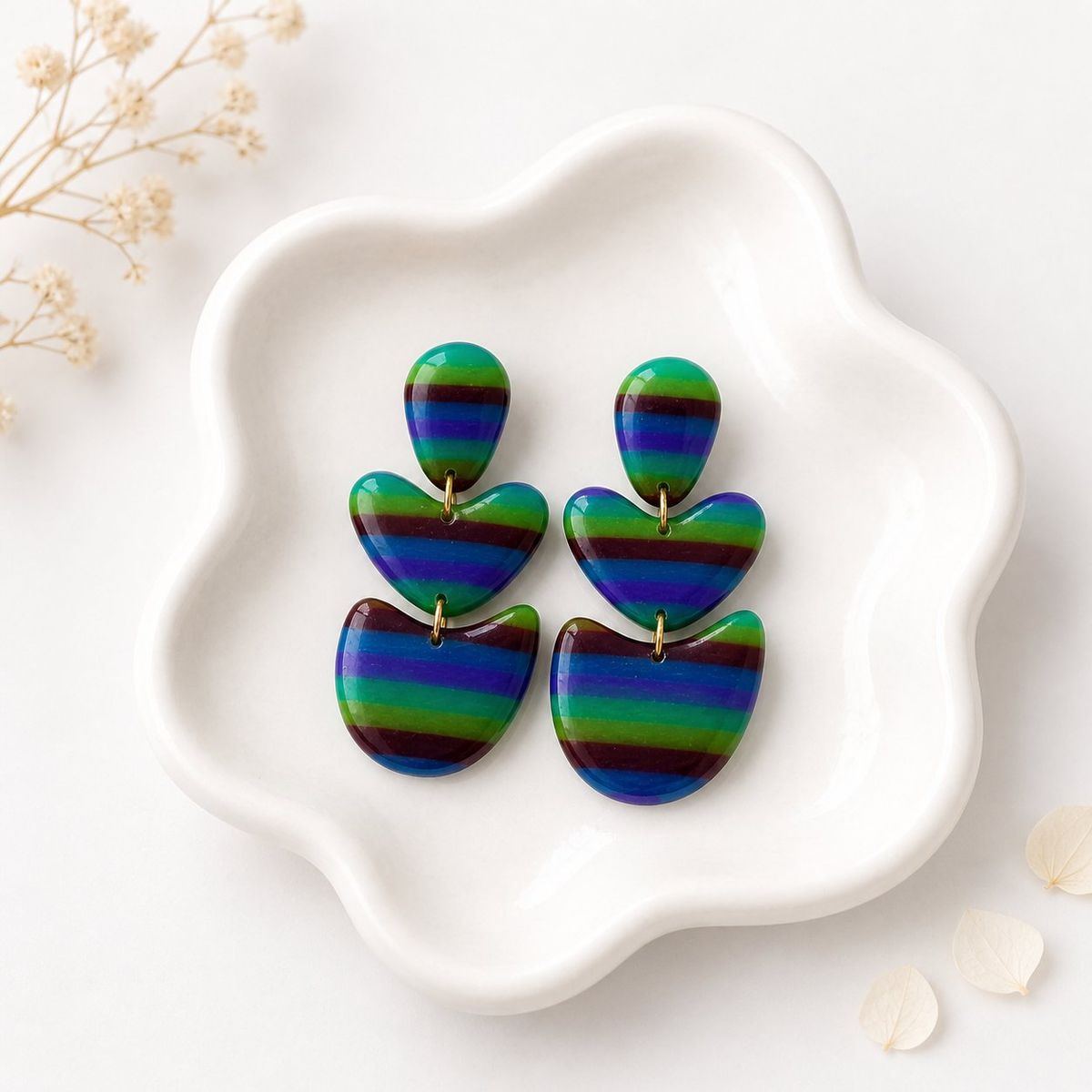 Three-tiered clay dangle earrings with horizontal green, blue, purple, and burgundy stripes, connected by small gold rings, displayed on a white ceramic dish.