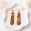 A pair of elongated teardrop dangle earrings with amber, rust, and gold glitter swirl patterns in translucent resin, attached to gold ball studs, displayed on a white ceramic dish.