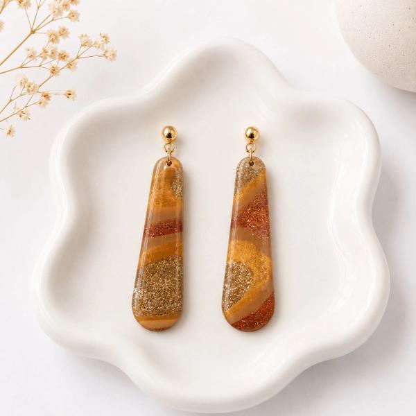 A pair of elongated teardrop dangle earrings with amber, rust, and gold glitter swirl patterns in translucent resin, attached to gold ball studs, displayed on a white ceramic dish.