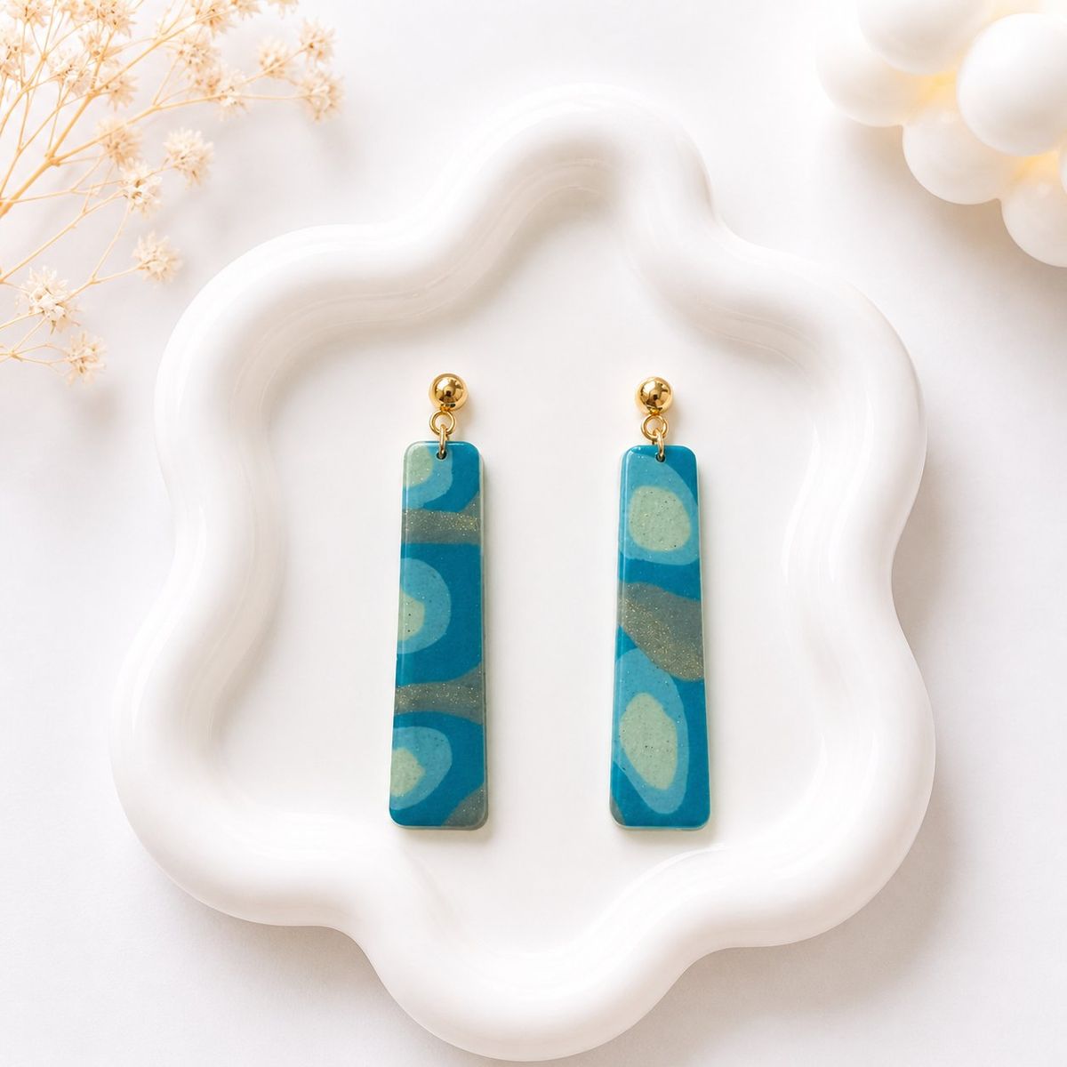 Two long rectangular clay dangle earrings with teal and sage green swirl pattern and gold glitter, hanging from gold ball studs on a white ceramic tray.