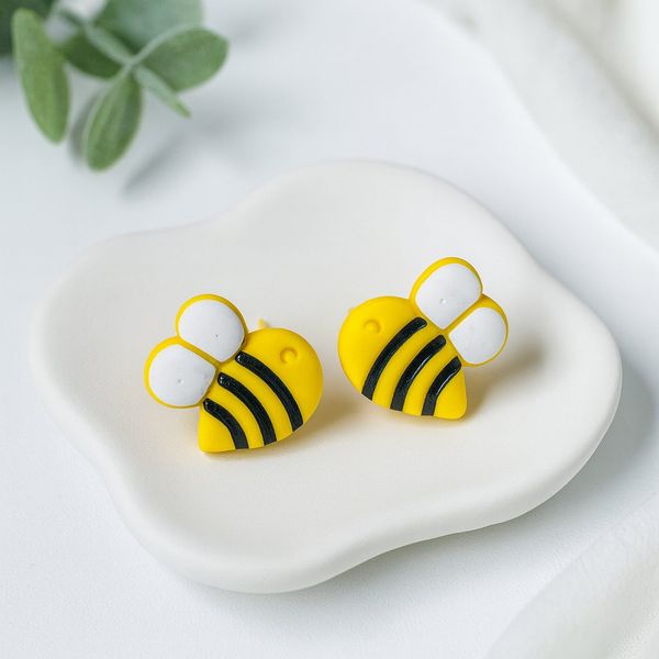 A pair of yellow and black striped bumblebee-shaped polymer clay stud earrings with white wings, displayed on a white ceramic dish.