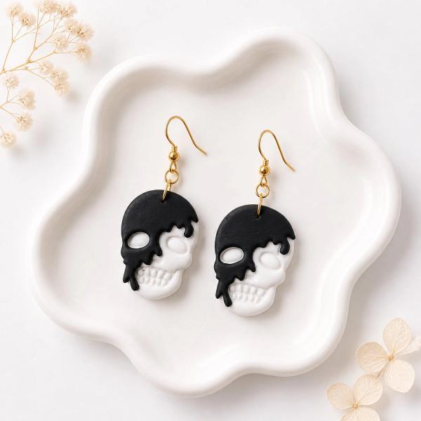 A pair of black and white clay skull dangle earrings with gold hooks, displayed on a white ceramic tray.