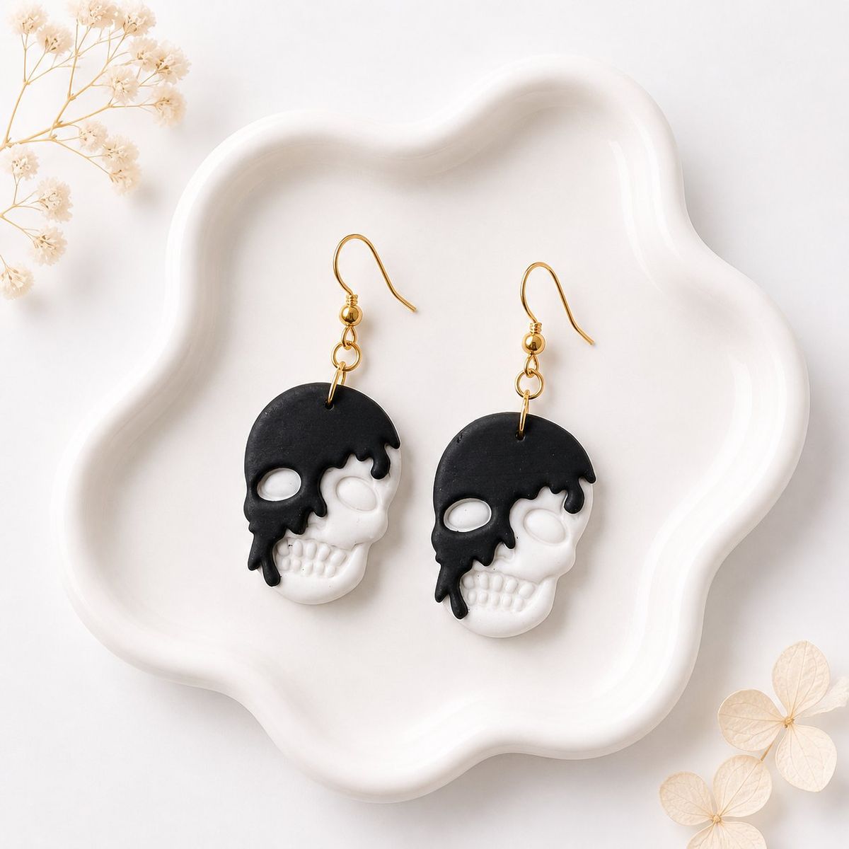 A pair of black and white clay skull dangle earrings with gold hooks, displayed on a white ceramic tray.