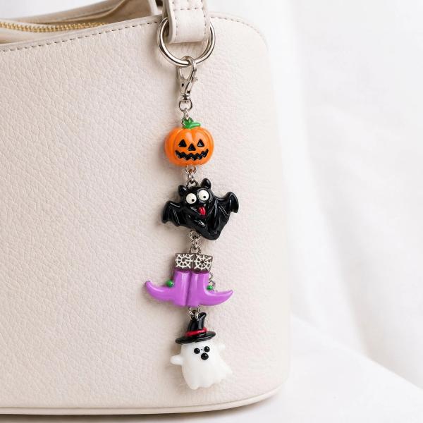 A Halloween-themed bag charm with four stacked resin figures — a jack-o'-lantern, black bat, purple witch hat, and white ghost with a top hat — attached to a cream handbag via a silver lobster clasp.