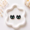 A pair of small black polymer clay cat face stud earrings with teal green eyes, displayed on a white wavy ceramic tray.