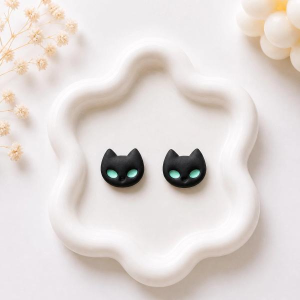 A pair of small black polymer clay cat face stud earrings with teal green eyes, displayed on a white wavy ceramic tray.