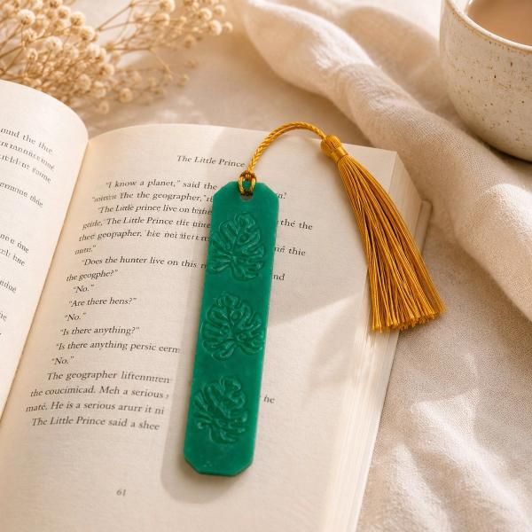 A teal green polymer clay bookmark with three embossed monstera leaf designs and a gold tassel, resting open on a book.