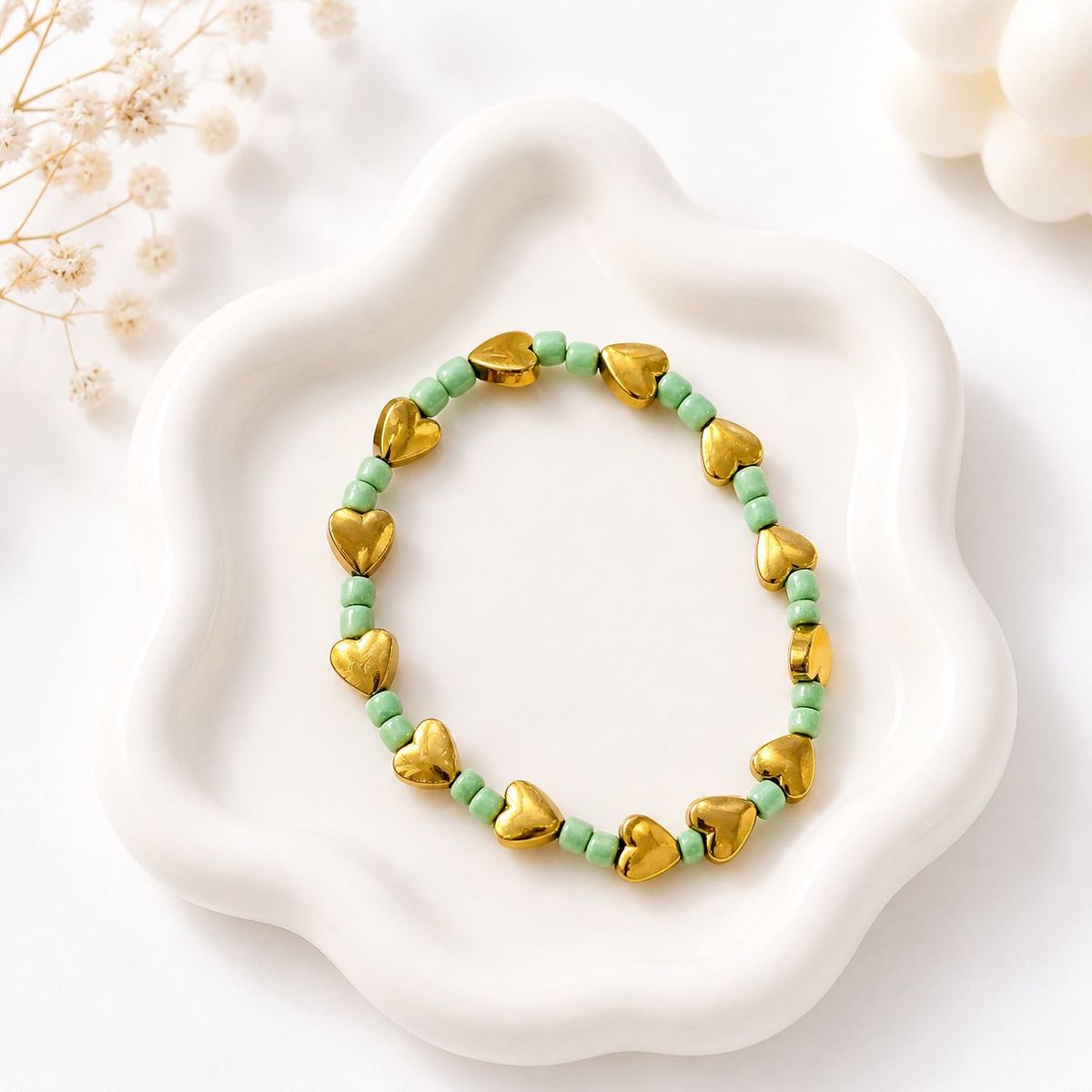 A stretch bracelet with alternating shiny gold-toned heart beads and small mint green seed beads, displayed on a white ceramic dish.