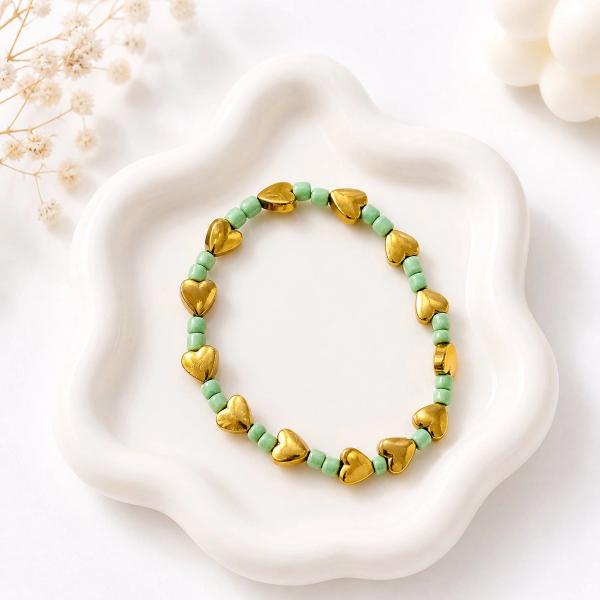 A stretch bracelet with alternating shiny gold-toned heart beads and small mint green seed beads, displayed on a white ceramic dish.