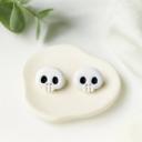A pair of white clay skull stud earrings with black eye sockets and small teeth, displayed on a cream ceramic dish.