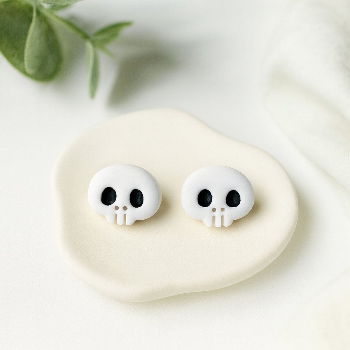 A pair of white clay skull stud earrings with black eye sockets and small teeth, displayed on a cream ceramic dish.