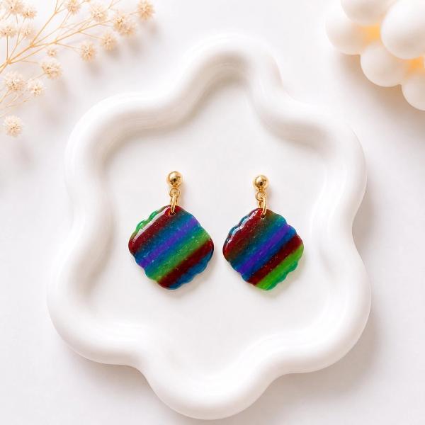 A pair of scallop-shaped clay dangle earrings with multicolor diagonal stripes in red, purple, blue, and green, hanging from gold ball studs on a white ceramic tray.