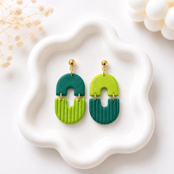 A pair of handmade clay dangle earrings with a two-part arch shape, each earring combining teal and lime green sections with vertical ribbed texture and gold-tone ball stud tops, displayed on a white ceramic tray.