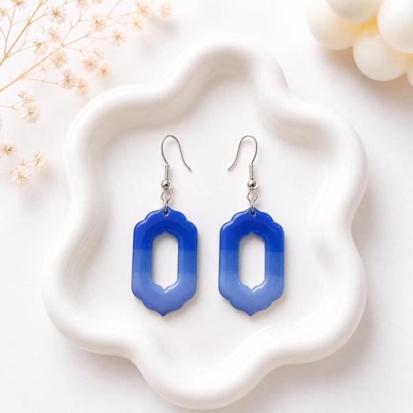 A pair of blue ombre open-frame clay dangle earrings with scalloped edges and silver fish hook findings, displayed on a white wavy dish.