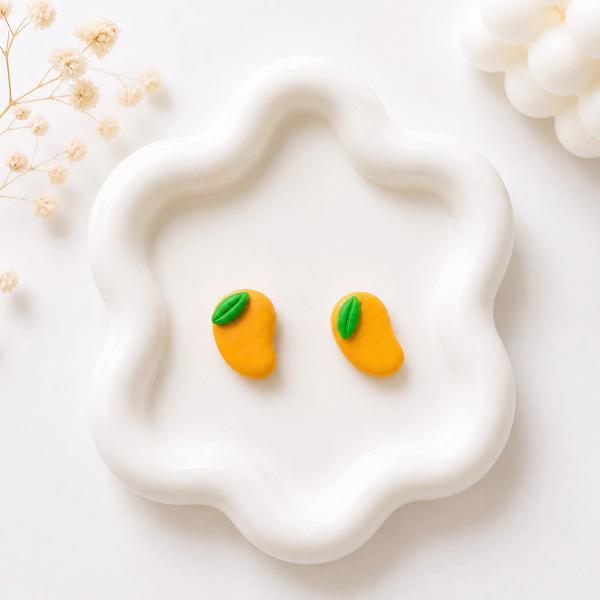 A pair of small orange mango-shaped polymer clay stud earrings with green leaf details, displayed on a white wavy ceramic tray.