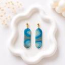 A pair of elongated clay dangle earrings with a blue and sage green wave pattern, hanging from gold ball studs, displayed on a white ceramic tray.