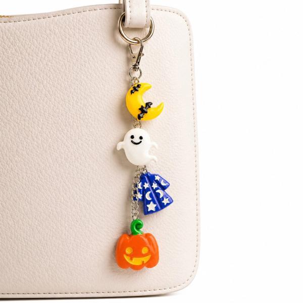 A Halloween-themed keychain with four resin charms — a crescent moon with bats, a white smiling ghost, a blue wizard hat with stars and moons, and an orange jack-o'-lantern — hanging from a silver lobster clasp on a cream handbag.