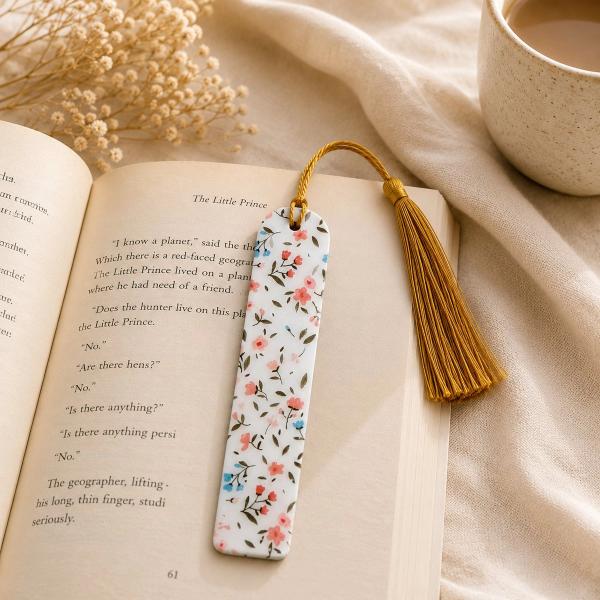 A slim white bookmark with a pink and blue ditsy floral pattern and olive green leaves, attached to a gold tassel via a looped cord, resting on an open book.