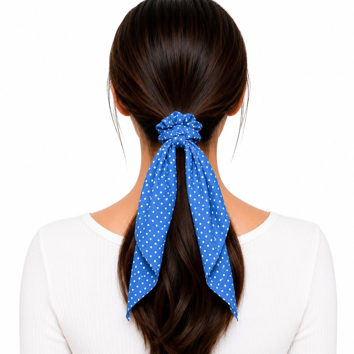 Back view of a woman wearing a cobalt blue and white polka dot scarf scrunchie holding a low ponytail, with two long fabric tails draping down.