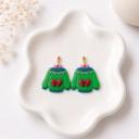 A pair of handmade polymer clay dangle earrings shaped like green Christmas sweaters with red bows, blue trim, and a geometric triangle neckline pattern, displayed on a white ceramic dish.