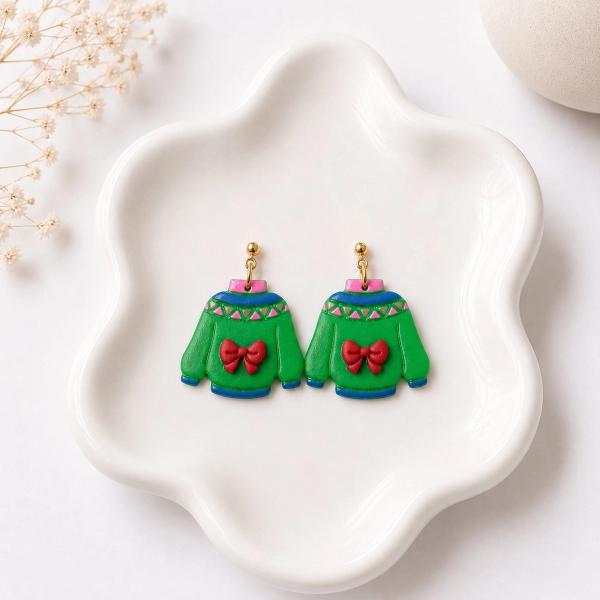 A pair of handmade polymer clay dangle earrings shaped like green Christmas sweaters with red bows, blue trim, and a geometric triangle neckline pattern, displayed on a white ceramic dish.