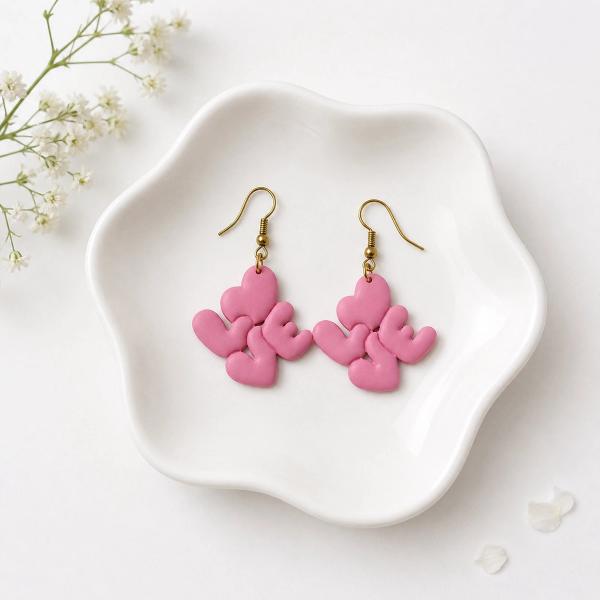 A pair of pink polymer clay dangle earrings spelling 'LOVE' in bubble heart-shaped letters on gold-tone fish hook wires, displayed on a white ceramic dish.