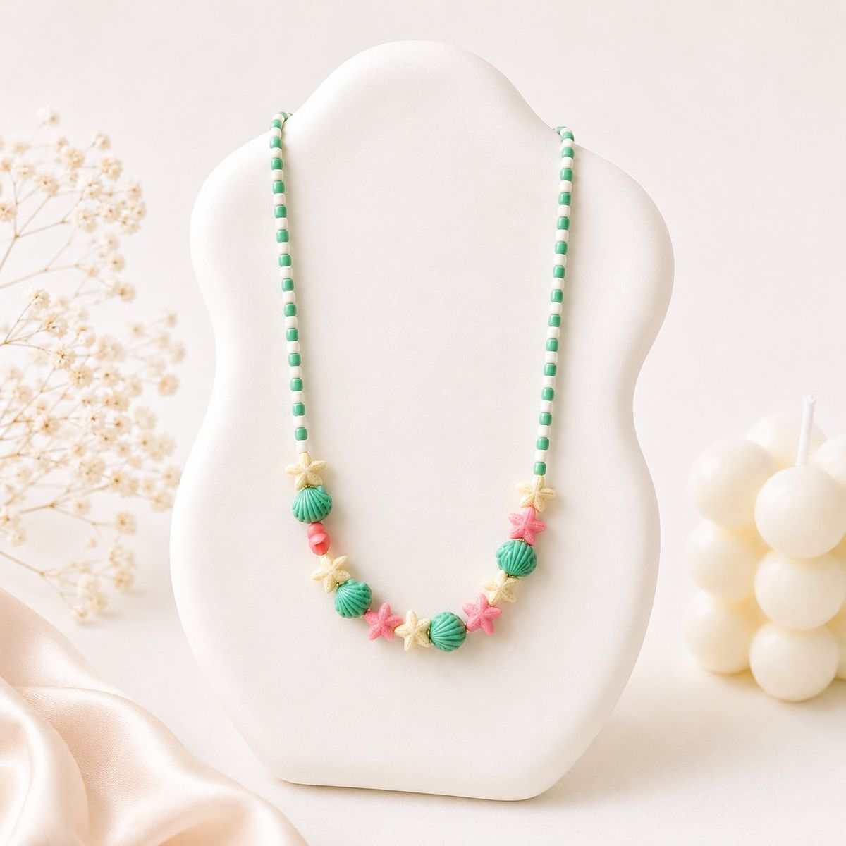 A beaded necklace displayed on a white bust featuring green clay seashell beads, pink and cream starfish charms, and alternating green and white seed beads.