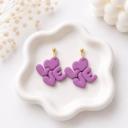 A pair of purple polymer clay dangle earrings shaped to spell LOVE with a heart, hanging from gold ball studs, displayed on a white ceramic dish.