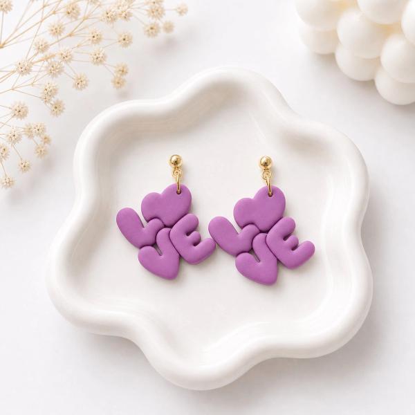 A pair of purple polymer clay dangle earrings shaped to spell LOVE with a heart, hanging from gold ball studs, displayed on a white ceramic dish.