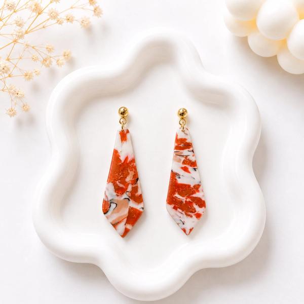 A pair of elongated kite-shaped clay dangle earrings with a red, white, and black marble pattern and gold flecks, hanging from gold ball studs on a white ceramic tray.