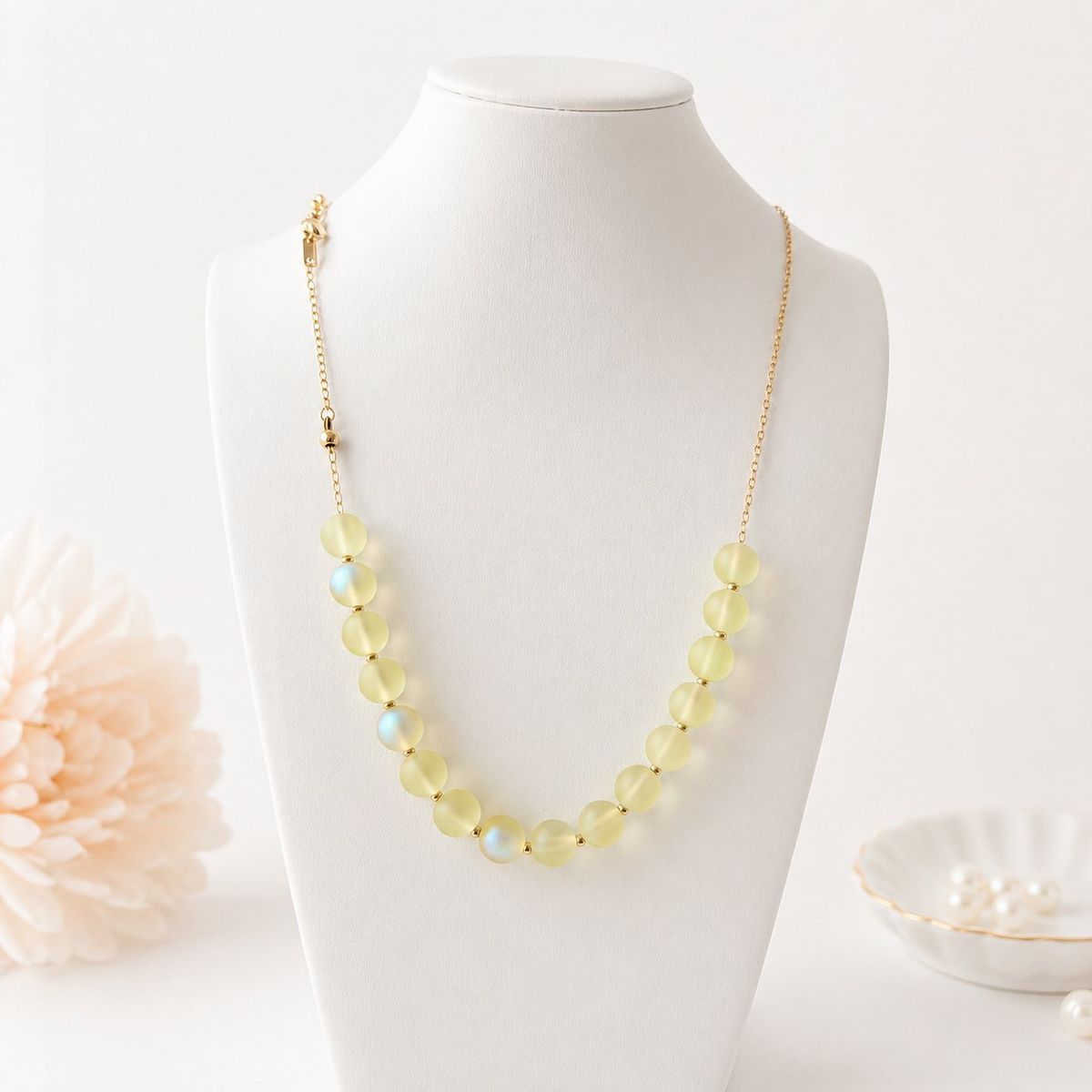 A necklace with matte frosted yellow-green round beads and small iridescent accent beads separated by gold-tone spacers, displayed on a white jewelry bust.