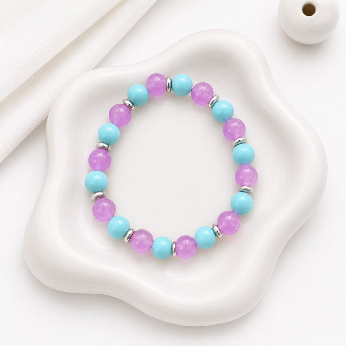 A stretch bracelet with alternating purple and teal round beads separated by small silver disc spacers, displayed on a white ceramic tray.
