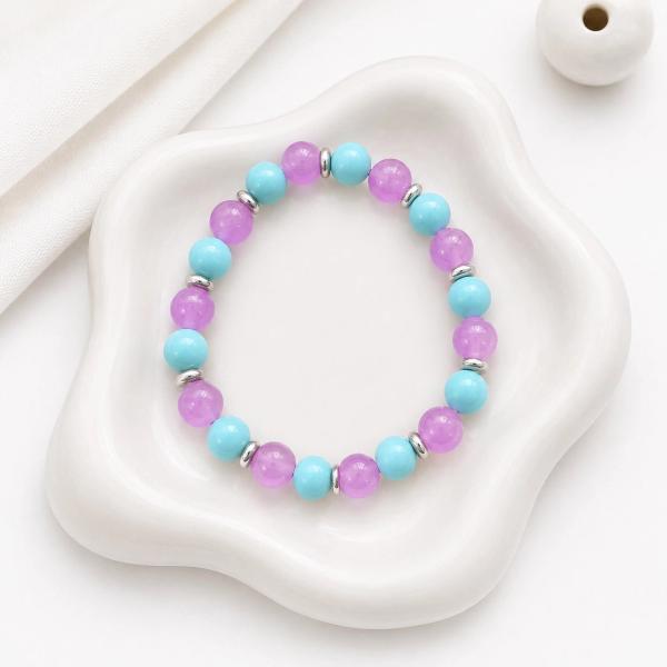 A stretch bracelet with alternating purple and teal round beads separated by small silver disc spacers, displayed on a white ceramic tray.