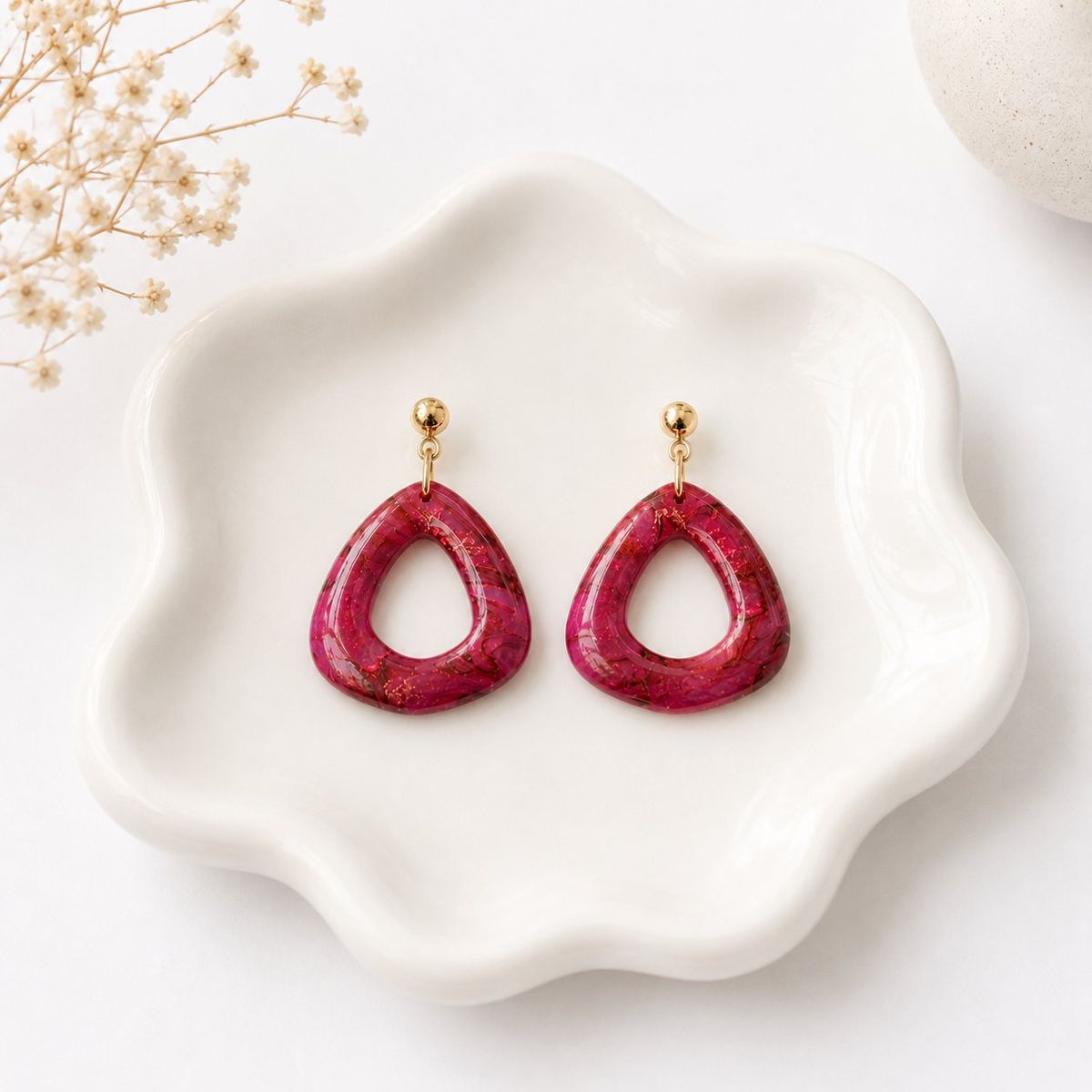 A pair of deep crimson marbled clay open teardrop dangle earrings with gold flecks, hanging from gold ball studs, displayed on a white scalloped dish.