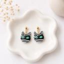 A pair of handmade polymer clay dangle earrings shaped like mummy cat faces with black and white stripes and teal green eyes, hanging from gold ball studs on small chain links, displayed on a white ceramic dish.