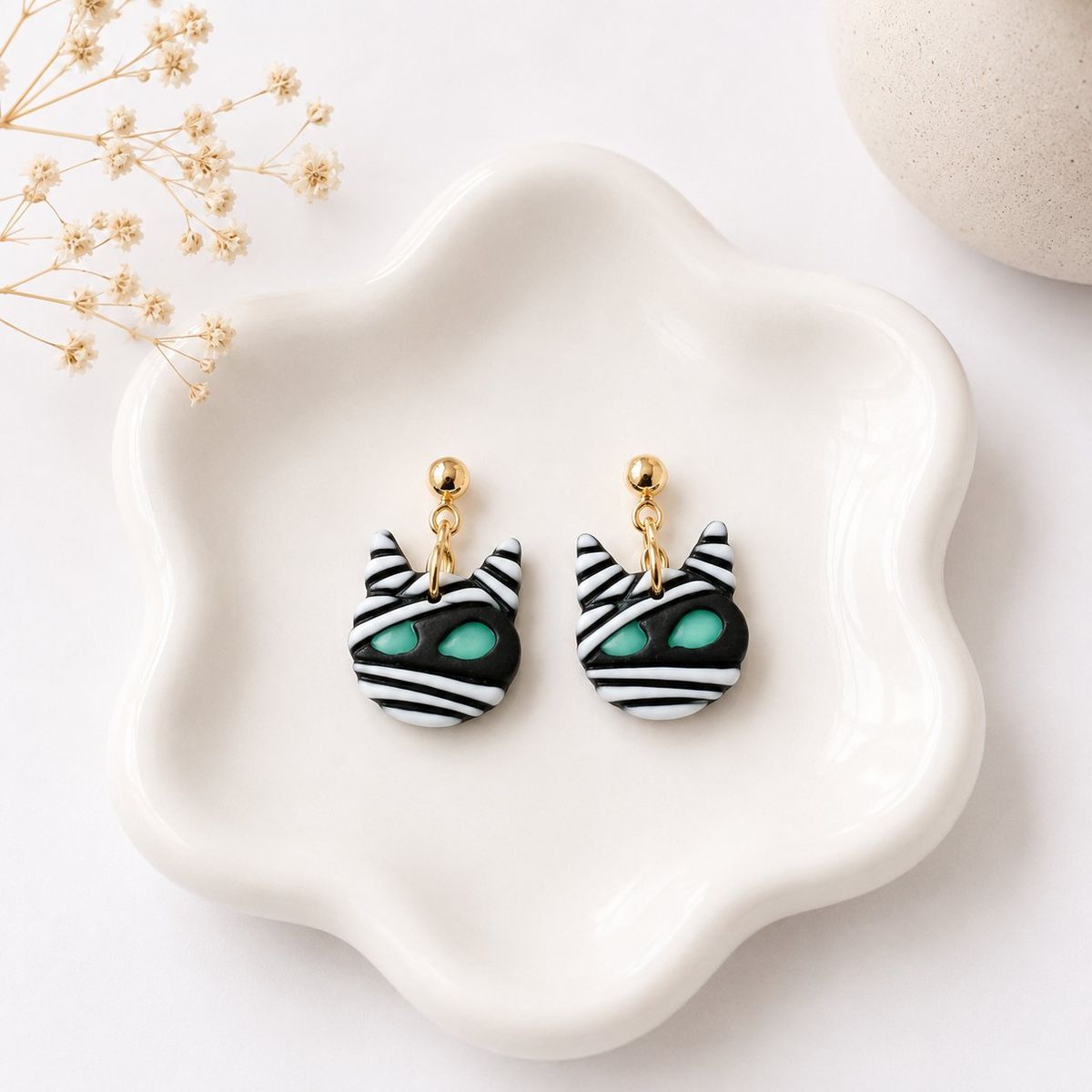 A pair of handmade polymer clay dangle earrings shaped like mummy cat faces with black and white stripes and teal green eyes, hanging from gold ball studs on small chain links, displayed on a white ceramic dish.