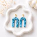 A pair of blue marbled arch-shaped clay dangle earrings with gold ball stud tops, displayed on a white ceramic tray.