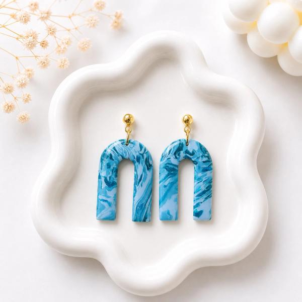 A pair of blue marbled arch-shaped clay dangle earrings with gold ball stud tops, displayed on a white ceramic tray.