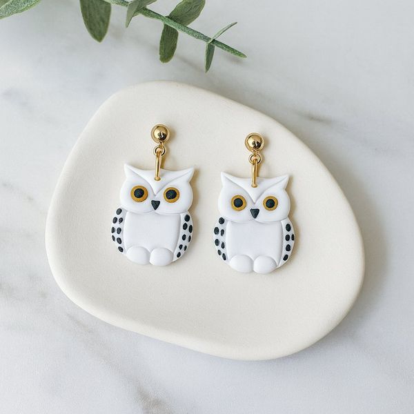 A pair of white polymer clay owl-shaped dangle earrings with black dot detailing, gold eyes, and gold ball studs, displayed on a cream clay tray against a marble background.