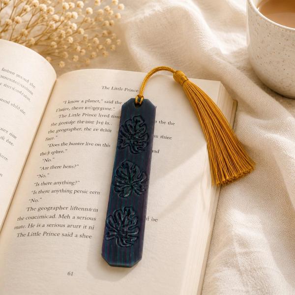 A dark navy polymer clay bookmark with embossed monstera leaf designs and a gold tassel, resting on an open book beside a cup of coffee.