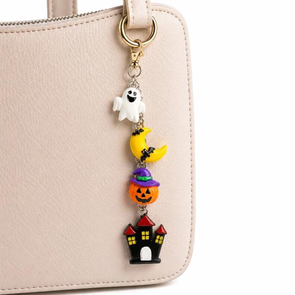 A Halloween-themed bag charm with four charms — a white ghost, yellow crescent moon with bat, orange jack-o'-lantern with purple witch hat, and black haunted house — hanging from a gold lobster clasp on a beige handbag.