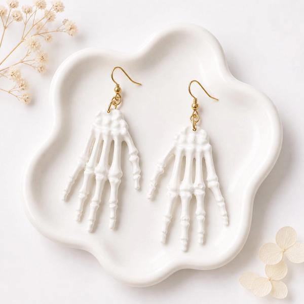 A pair of white clay skeleton hand dangle earrings with gold fishhook hardware displayed on a white ceramic tray.