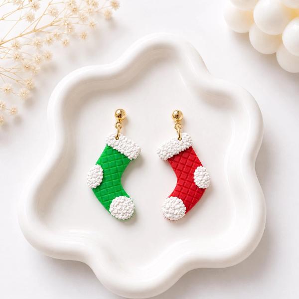 Two mismatched polymer clay Christmas stocking dangle earrings — one green and one red — with white textured trim and gold ball stud tops, displayed on a white ceramic tray.